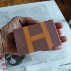 Savon H artisanal tenu en main sur Wimzzi, savon solide graphique aux teintes terracotta et mauve, soin fait main original, cadeau beauté utile pour peau douce, salle de bain créative et routine naturelle / Handgemaakte H zeep in de hand op Wimzzi, grafische vaste zeep in terracotta en mauve, originele verzorging en nuttig beautycadeau voor zachte huid en natuurlijke routine / Handmade H soap held in hand on Wimzzi, graphic solid soap in terracotta and mauve tones, original care and useful beauty gift for soft skin and natural routines